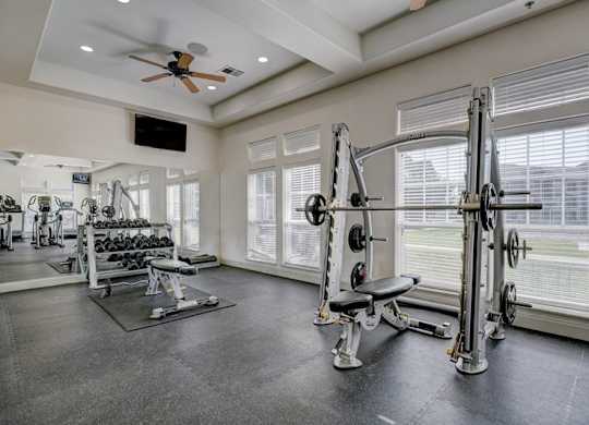 A fitness center here at Liberty Pointe featuring a weight training area with a squat rack, adjustable bench, and free weights. Large windows and mirrors create a bright, open workout space with a clean and modern feel.