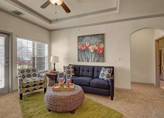 Here at Liberty Pointe, a cozy living area with a deep blue sofa and a patterned accent chair paired with a bold green rug. A woven round ottoman coffee table sits centered with tray décor and candles, while a tulip wall art piece adds warm color above the sofa and natural light comes through the window and patio door.