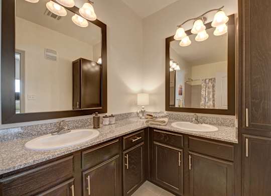 Here at Liberty Pointe, a bright bathroom vanity area with granite countertops and dark wood cabinetry, featuring dual white sinks and large framed mirrors with warm vanity lighting. Simple decor like folded neutral towels, soap dispensers, and a small table lamp create a clean, polished, and welcoming feel.