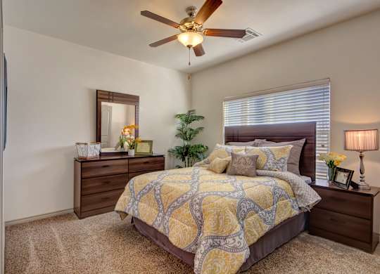 A bedroom here at Liberty Pointe featuring soft neutral walls, plush carpet, and a bed with yellow and gray patterned bedding and layered neutral accent pillows. The space includes dark wood nightstands and a dresser. Includes a soft green plant in the corner of the space with a wooden ceiling fan and fan light, creating a cozy atmosphere.