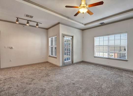 A living room here at Liberty Pointe featuring soft beige walls, plush neutral carpet, and bright natural light from multiple windows and a glass door. The space includes tray ceiling detail with a ceiling fan and modern track lighting, creating a bright, open, and comfortable atmosphere.