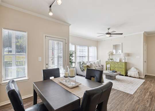 A dining and living area here at Lincoln at Central Park featuring a dark wood dining table with four black upholstered chairs, styled with a neutral table runner and glass candle centerpieces. The space opens into a bright living room with a light neutral sofa, a green accent chair, and a standout green decorative dresser that adds a fresh pop of color. Large windows bring in natural light.
