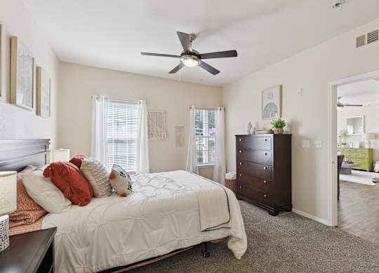 A bedroom here at Lincoln at Central Park featuring a dark wood bed styled with white bedding and standout rust-orange accent pillows and textured throw for warm contrast. Matching dark wood furniture including a tall dresser adds storage, while soft white curtains, neutral wall décor, and plush carpet create a bright, comfortable, and inviting space.