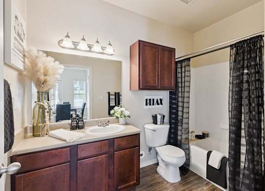 A bathroom space here at Lincoln at Central Park featuring a dark wood vanity, black shower curtain, bright mirror lighting, white tub and toilet, and warm neutral walls with clean, modern styling.