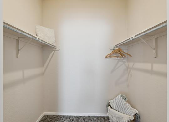 A walk-in closet here at Lincoln at Central Park featuring beige walls, carpet flooring, dual hanging rods, upper shelving, and a few wooden hangers with a light decorative pillow for a clean, organized look.