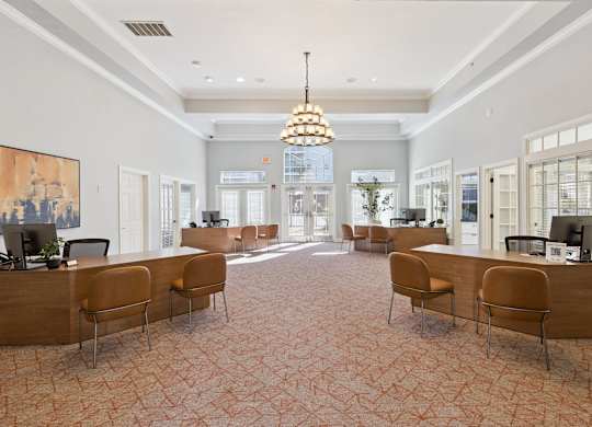 A welcoming leasing office lobby here at Lincoln at Central Park with wood desks, comfortable guest seating, and soft patterned carpet. A large chandelier and tall windows create a bright, upscale, and professional first impression for residents and visitors.
