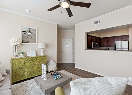 A living room here at Lincoln at Central Park featuring a bright, open layout with a light gray sofa, a green accent chair, and a green decorative dresser that adds a standout pop of color. The space includes a square coffee table on a light area rug, wood-style flooring, and a pass-through view into the kitchen with dark wood cabinets and stainless steel appliances.