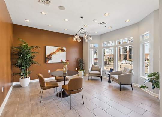 A stylish seating lounge here at Lincoln at Central Park with a warm accent wall, round table with chairs, and modern light fixture. A large corner window and comfortable armchairs create a bright, inviting space for relaxing or casual meetings.