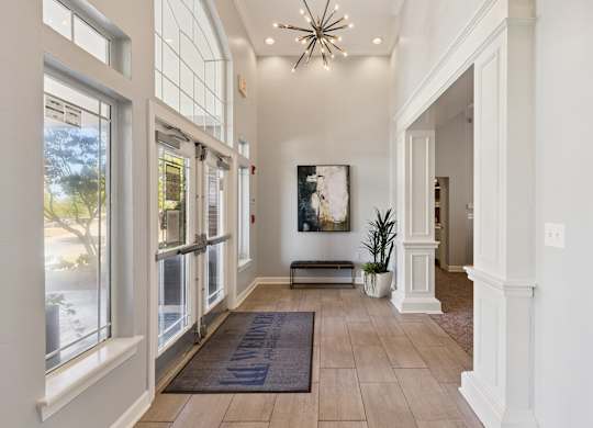 A bright entry lobby here at Lincoln at Central Park with tall windows, glass entry doors, and light wood-style flooring. A modern chandelier, decorative artwork, and greenery create a clean, welcoming first impression for residents and guests.