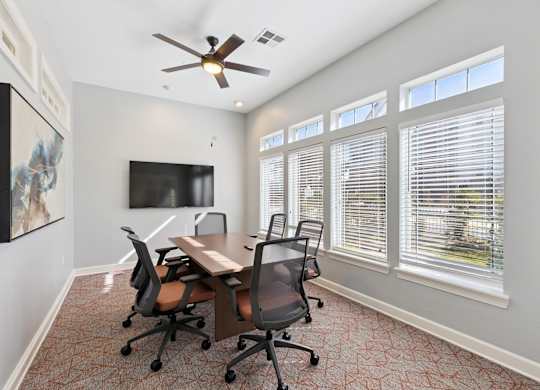 A private meeting room here at Lincoln at Central Park with a wood conference table, rolling office chairs, and a wall-mounted TV. A ceiling fan and large windows with blinds create a bright, comfortable workspace for meetings or remote work.
