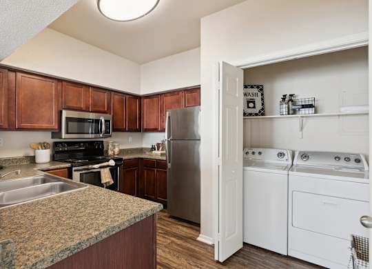 A kitchen here at Lincoln at Central Park featuring warm wood cabinets, granite-style countertops, and stainless steel appliances including a microwave, range, and refrigerator. The space opens to a convenient in-unit laundry area with a full-size washer and dryer, upper storage shelf, and simple organized laundry accessories, all set on wood-style flooring with bright overhead lighting.