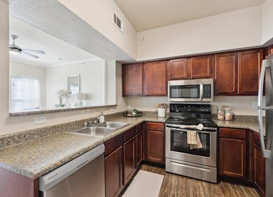 A kitchen here at Lincoln at Central Park featuring rich wood cabinetry, granite-style countertops, and stainless steel appliances including a microwave, range, dishwasher, and refrigerator. The space includes a double-basin sink with a pass-through opening to the living area, simple countertop accessories, and wood-style flooring, creating a bright and clean cooking space.