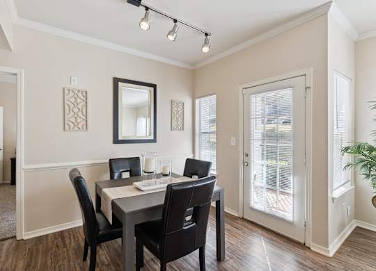 A dining area here at Lincoln at Central Park featuring a dark wood dining table paired with four black upholstered chairs, set on warm wood-style flooring. Natural light enters through a glass patio door and side window with white blinds, while a wall mirror, decorative wall panels, and a simple candle centerpiece create a clean, welcoming, and comfortable space.