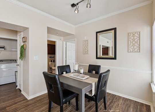 A dining area here at Lincoln at Central Park featuring a dark wood dining table with four black upholstered chairs, set on warm wood flooring. A neutral table runner and glass candle holders create a simple, welcoming centerpiece, while a wall mirror and decorative panels add visual interest.