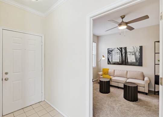 A entryway here at Quail Landing with a bright front door, clean tile flooring, and a view into the living area featuring a beige sofa, yellow throw blanket, neutral carpet, and soft overhead lighting.