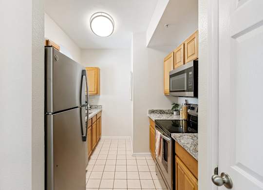 A compact kitchen here at Quail Landing with warm wood cabinets, granite-style countertops, stainless steel refrigerator and range, light tile flooring, and bright overhead lighting in a clean, efficient layout.