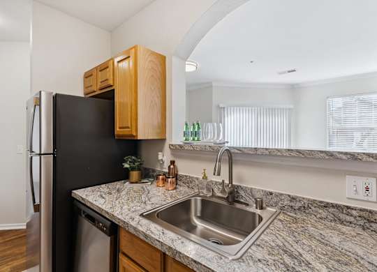 A kitchen here at Quail Landing featuring warm wood cabinets, granite-style countertops, a stainless steel sink and dishwasher, a pass-through opening, and bright natural light for a clean, inviting feel.