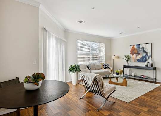 A living and dining area here at Quail Landing featuring warm wood floors, a cozy neutral sofa, a round dining table, soft area rug, indoor plants, and bright natural light from large windows and sliding doors.