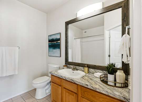 A bathroom here at Quail Landing with a granite vanity, wood cabinets, large framed mirror, white sink and toilet, neutral tile floor, simple wall art, folded towels, and soft lighting creating a clean, calm look.