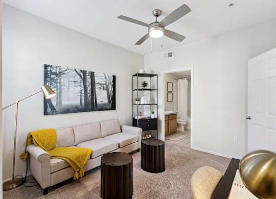 A home office here at Quail Landing with soft carpet flooring, a beige sofa accented by a mustard throw, black shelving, small round stools, a ceiling fan, and warm natural light creating a flexible, inviting workspace.