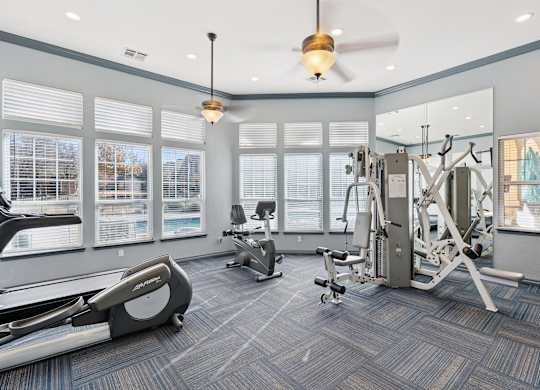 A fitness center here at Quail Landing with modern cardio and strength equipment, blue patterned flooring, ceiling fans, and large windows that bring in bright natural light.