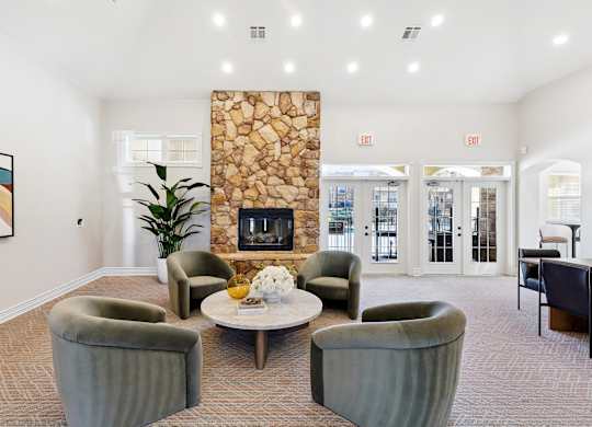 A community lounge here at Quail Landing with four soft green lounge chairs around a round coffee table, a tall stone fireplace, light carpet flooring, large windows, and bright recessed ceiling lighting.