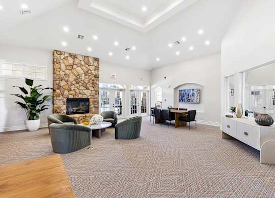 A community lounge here at Quail Landing with soft green lounge chairs, a round coffee table, a tall stone fireplace, light carpet flooring, and bright recessed ceiling lighting.