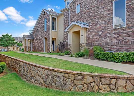 A community exterior walkway here at Quail Landing with brick apartment buildings, stone retaining wall, green landscaping, covered entryways, and a clean concrete path under a bright blue sky.
