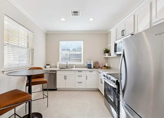 A kitchen here at Quail Landing with white cabinets, stainless steel appliances, light tile flooring, a small round dining table with brown leather stools, bright windows with blinds, and a clean, modern layout.