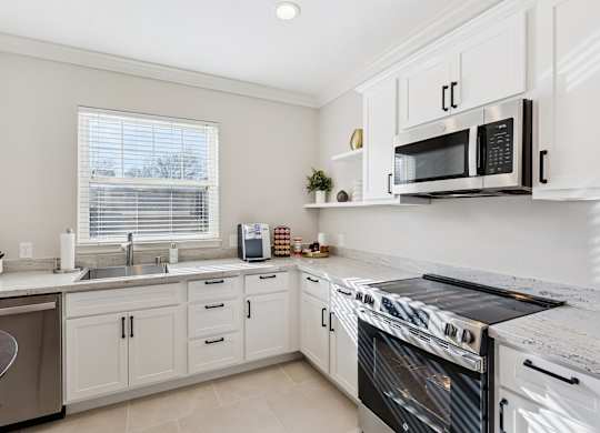 A kitchen here at Quail Landing with bright white cabinets, stainless steel appliances, light countertops, soft tile flooring, open shelving with greenery, and a sunlit window above the sink.