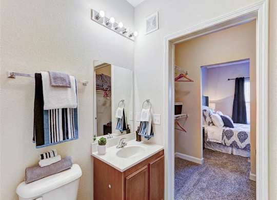 A bathroom with a toilet, sink, mirror, and a bedroom in the background.