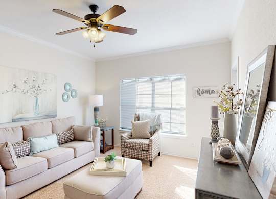 A bright living room here at Stoneleigh on May with beige carpet, a soft neutral sofa, a patterned accent chair, and a ceiling fan. Sunlight filters through wide windows, creating a warm, inviting, and comfortable atmosphere.