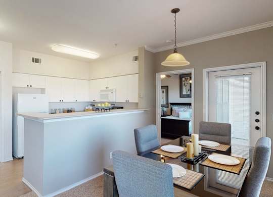 A welcoming dining area here at Stoneleigh on May with a glass-top table, upholstered chairs, and pendant lighting. A breakfast bar connects to the kitchen, while a sliding glass door with blinds adds natural light and an open feel.