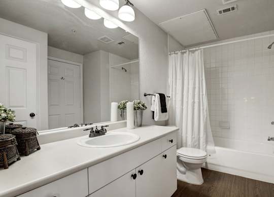 A clean bathroom here at Stoneleigh on May with a wide vanity, white cabinets, and a large mirror under bright lighting. A bathtub-shower combo with white tile and curtain creates a fresh, simple, and functional space.