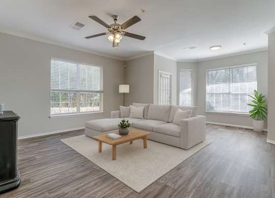 A bright living room here at Sycamore Farms with wood-style flooring, a neutral sectional sofa, and a ceiling fan with light. Large windows with blinds bring in natural light, creating an open, calm, and inviting atmosphere.
