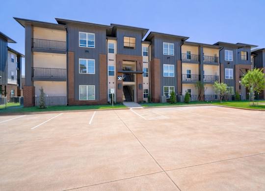 A apartment building exterior here at The Encore with modern three-story architecture, gray and brick accents, private balconies, and landscaped grounds under a clear blue sky.