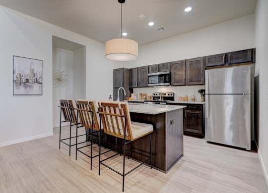 A kitchen here at The Encore with a spacious island, dark wood cabinetry, and stainless steel appliances. Light flooring, a pendant light, and wood-tone bar stools create a warm, modern gathering space.