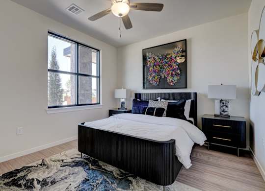 A bedroom here at The Encore with a black bed frame, crisp white bedding, and colorful wall art above the headboard. Light flooring, matching nightstands with lamps, a ceiling fan, and a large window create a bright, modern feel.