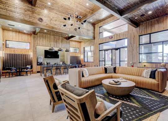 A clubhouse lounge here at The Encore with a tan sectional sofa, round wood coffee table, and warm wood-paneled ceilings. The open space feels inviting and stylish, with cozy seating areas, modern lighting, and large windows for natural light.