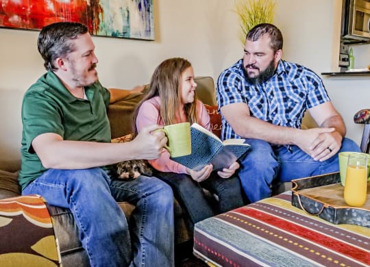 Two dads sitting on the couch reading with their daughter in their living room at Bristol Square Apartments