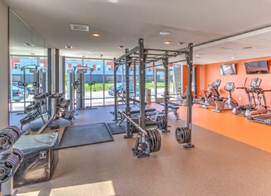 Bright gym with large windows, weightlifting equipment, and exercise machines. Orange accent wall adds warmth at Stitchweld Apartments.