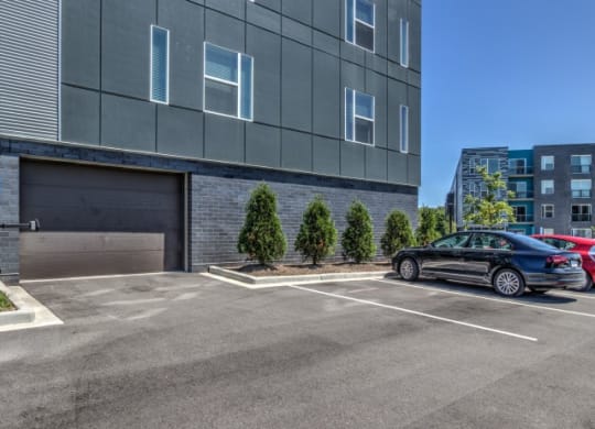 Parking lot with gray building on the left, featuring small trees lined along the side. A parked black car is visible under a clear blue sky at Stitchweld Apartments.