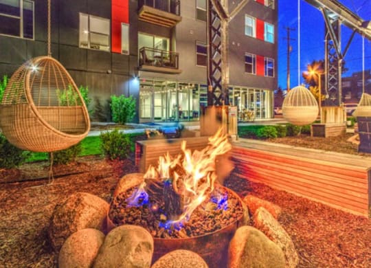 A cozy outdoor fire pit surrounded by stones, set in a modern apartment courtyard with colorful lighting and hanging chairs, creating a relaxed ambiance at Stitchweld Apartments.