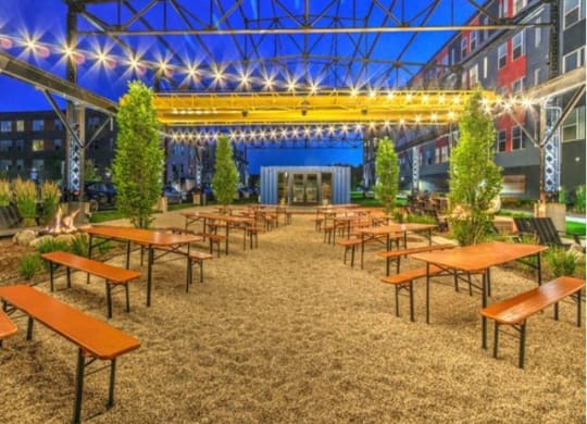 Outdoor garden at dusk with empty wooden tables and benches on gravel. Strings of lights hang above, creating a cozy, inviting atmosphere at Stitchweld Apartments.