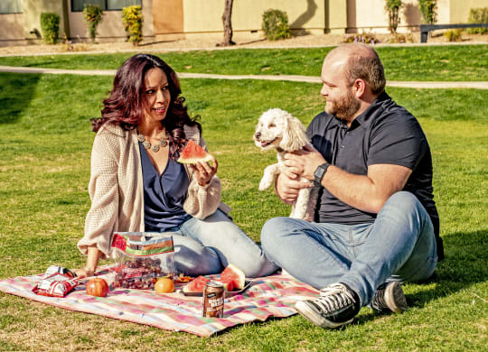 A couple enjoys a picnic on a sunny day, sitting on a blanket with snacks and watermelon. The woman holds a slice while the man smiles at a small dog. The Alaskan Apartments.
