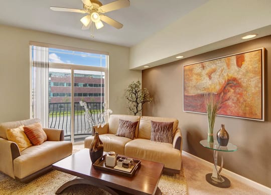 Bright modern living room here at Park Plaza 2 Apartments with beige sofas, abstract painting, ceiling fan, and large window showing a view of an office building. Cozy and inviting.