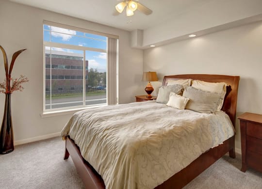 Modern bedroom here at Park Plaza 2 Apartments with a large window view, featuring a wooden bed with beige and gray bedding, side tables, a tall vase, and a warm, inviting ambiance.