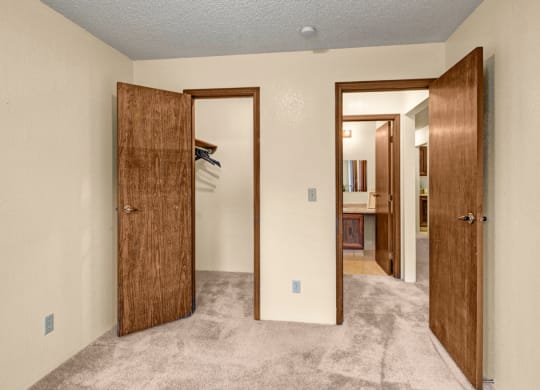 A small bedroom here at Park Plaza Apartments with beige walls and carpet. Two open wooden doors show a closet with hangers on the left and a bathroom with a mirror on the right.
