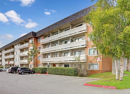 Four-story Park Plaza apartment building with white balconies, surrounded by green trees and a parking lot with several cars, under a clear blue sky.
