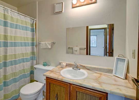 A bathroom here at Park Plaza Apartments with a striped shower curtain in green, blue, and white. A marble sink counter holds soap and a decorative sign. Bright, clean ambiance.